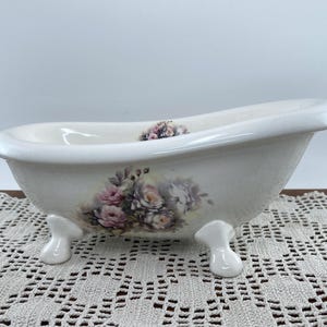 May include: A white ceramic bathtub with floral accents. The tub has a classic clawfoot design and is decorated with pink and purple roses. The tub is sitting on a white lace tablecloth.