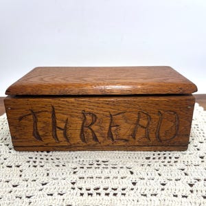 May include: A rectangular wooden box with a medium brown finish and visible wood grain. The front panel features the word THREAD carved into the surface in capital letters. The box has a flat, hinged lid that sits flush with the base.