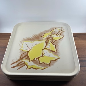 May include: A square, off-white serving tray with a floral design. The tray features a cluster of yellow leaves and brown stems against a light brown background. The tray has a slightly raised edge and is stacked with other trays.