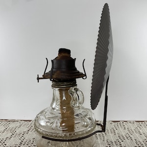 May include: A vintage oil lamp with a clear glass reservoir and a decorative floral pattern. The lamp has a metal base and a dark brown top. A dark gray fan-shaped reflector is attached to the side.