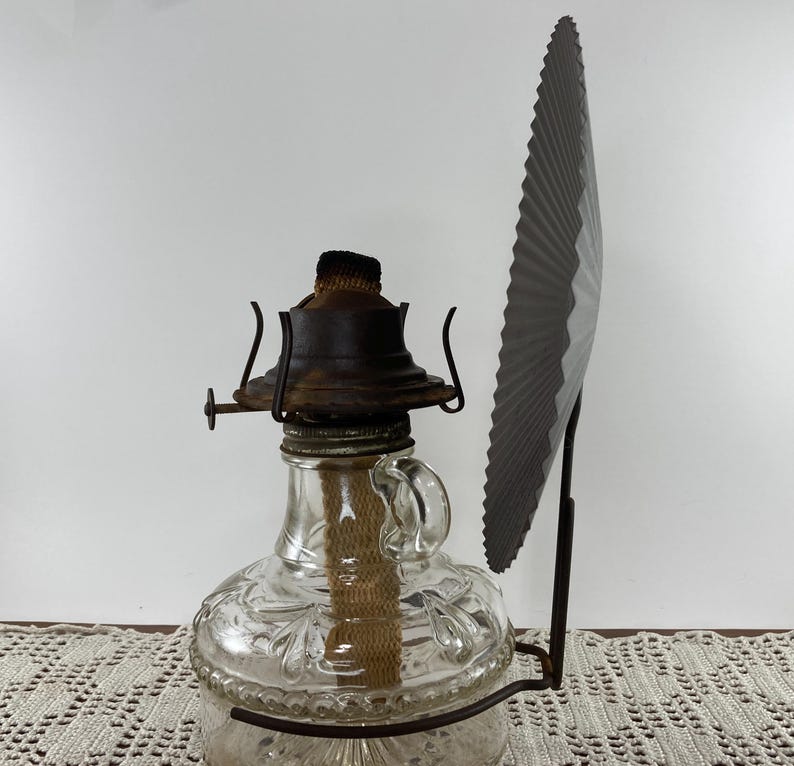 May include: A vintage oil lamp with a clear glass reservoir and a decorative floral pattern. The lamp has a metal base and a dark brown top. A dark gray fan-shaped reflector is attached to the side.