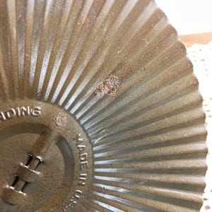 May include: Close-up of a vintage, circular, metal baking pan with a fluted design. The pan has a distressed, aged appearance with visible rust and wear. The text "MADE IN U.S.A." is stamped on the side.