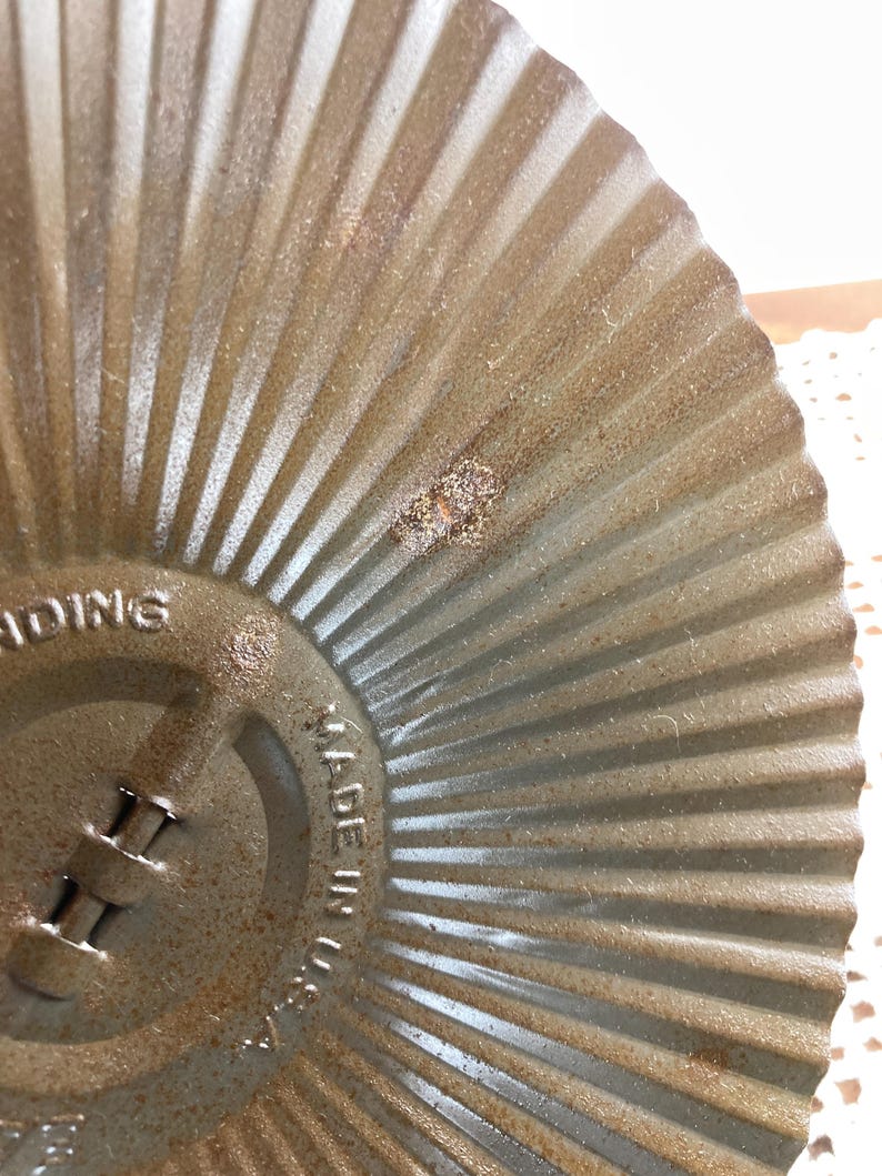 May include: Close-up of a vintage, circular, metal baking pan with a fluted design. The pan has a distressed, aged appearance with visible rust and wear. The text "MADE IN U.S.A." is stamped on the side.