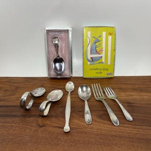 May include: A collection of silver-toned flatware including two curved baby spoons, a long-handled spoon, a decorative spoon, and two forks. One spoon is in a clear plastic box, and a yellow box features a kangaroo illustration and the text "something shiny inside."