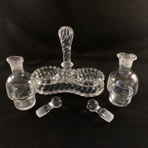 May include: A clear glass salt and pepper shaker set with a central handle. The set has two small bowls for salt and pepper and two larger bottles for oil and vinegar. The set is made of clear glass with a textured design.