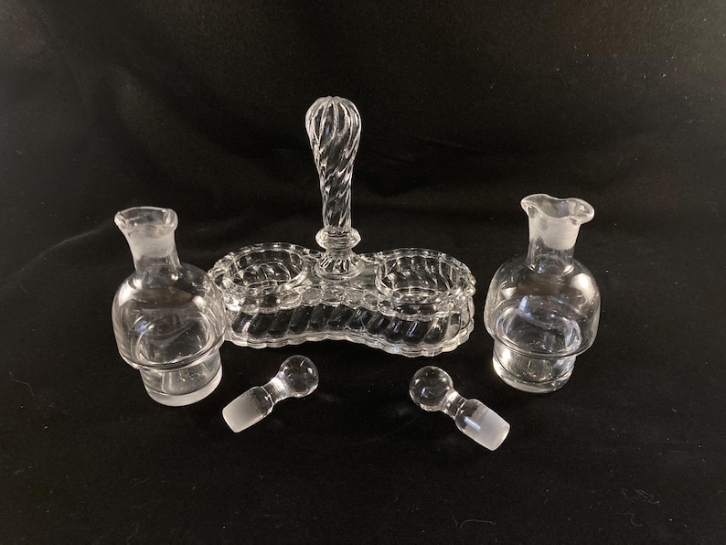 May include: A clear glass salt and pepper shaker set with a central handle. The set has two small bowls for salt and pepper and two larger bottles for oil and vinegar. The set is made of clear glass with a textured design.