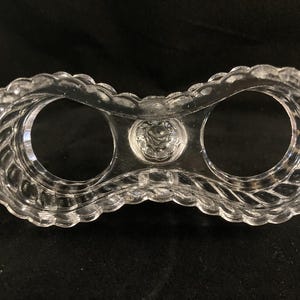 May include: A clear glass egg holder with two compartments. The holder has a decorative, twisted design around the rim.