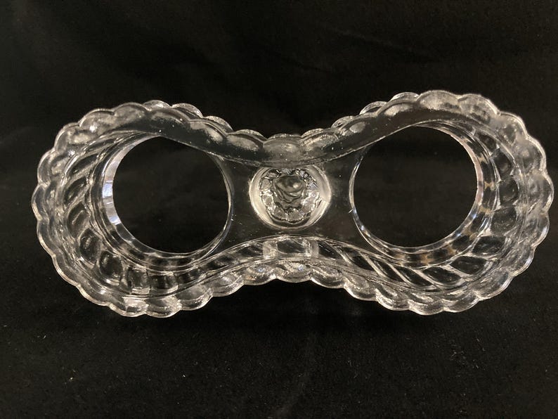 May include: A clear glass egg holder with two compartments. The holder has a decorative, twisted design around the rim.