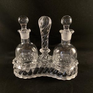 May include: A clear glass oil and vinegar cruet set with three bottles and a twisted handle. The bottles have a round body and a small opening at the top. The cruet set is on a clear glass base with a decorative design.