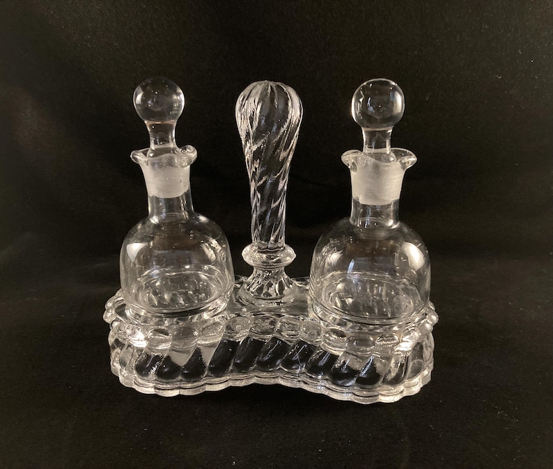 May include: A clear glass oil and vinegar cruet set with three bottles and a twisted handle. The bottles have a round body and a small opening at the top. The cruet set is on a clear glass base with a decorative design.