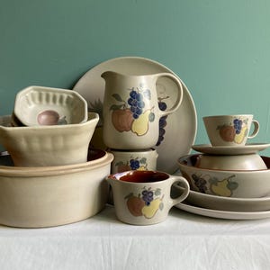 May include: A collection of beige ceramic tableware with fruit designs. Includes a pitcher, bowls, plates, and cups. The fruit designs feature grapes, apples, and pears in shades of blue, red, and yellow.