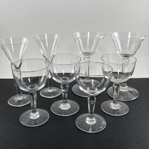 May include: A collection of clear glass stemware, including coupe, champagne, and martini glasses. The glasses have various bowl shapes and stem designs, arranged on a dark surface. The glassware is suitable for serving cocktails and beverages.