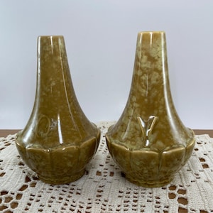 May include: Two vintage ceramic vases with a speckled, olive-green glaze. The vases have a flared base with a scalloped edge and a tall, slightly tapered neck. The vases are displayed on a cream-colored lace doily.