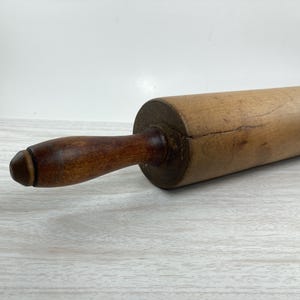 May include: A wooden rolling pin with a dark brown handle and a light brown rolling surface. The rolling pin is made of wood and is designed for rolling out dough. The handle is attached to the rolling surface.