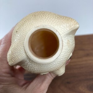 May include: A vintage, off-white piggy bank with a textured surface. The bank has a large opening on top and a small eye detail. The bottom of the bank is not visible. A red lid is visible in the bottom right corner.