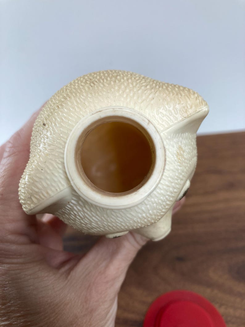 May include: A vintage, off-white piggy bank with a textured surface. The bank has a large opening on top and a small eye detail. The bottom of the bank is not visible. A red lid is visible in the bottom right corner.