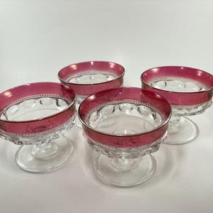 May include: Four clear glass dessert dishes with a ruby red rim and base. The bowls have a decorative pattern around the rim and base. The dishes are on a white background.