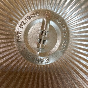May include: Close-up of a vintage metal pie pan. The center features the text "PAT. PENDING", "MADE IN USA", and "EAGLE". The pan has a ridged design radiating from the center.