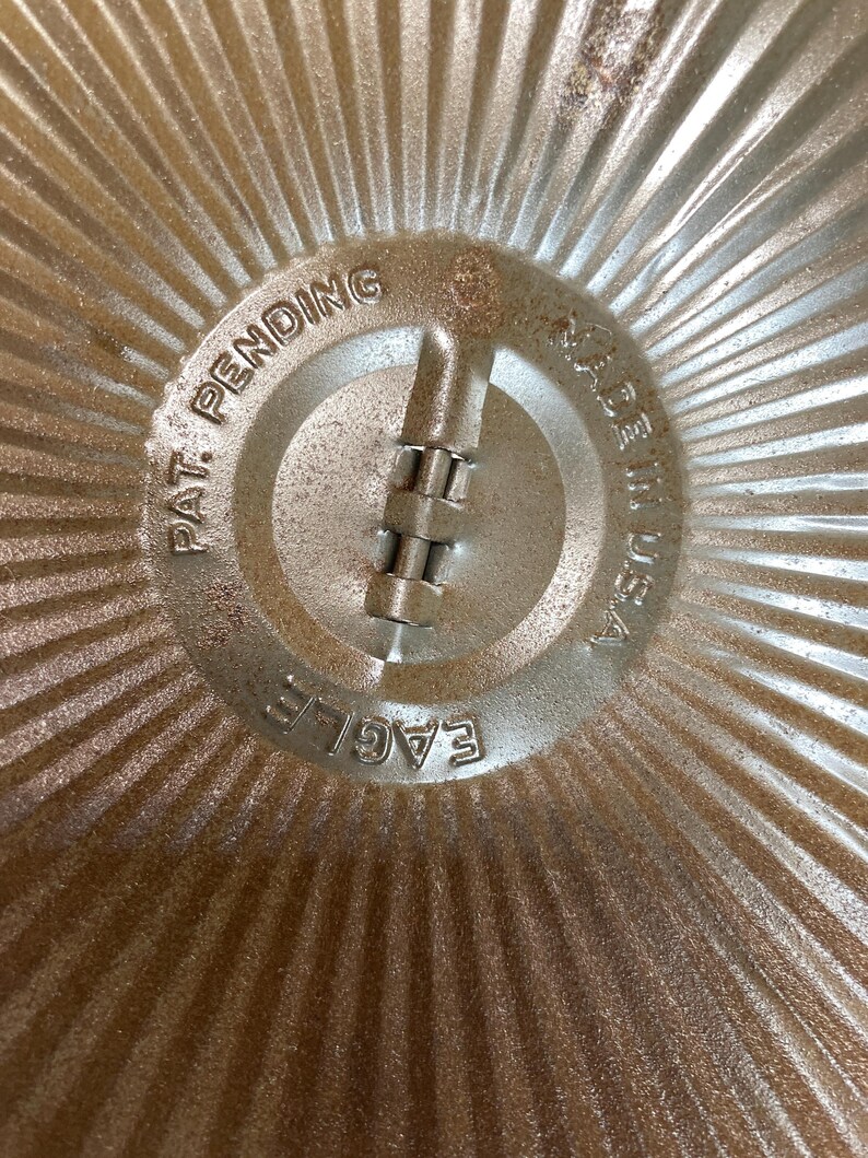 May include: Close-up of a vintage metal pie pan. The center features the text "PAT. PENDING", "MADE IN USA", and "EAGLE". The pan has a ridged design radiating from the center.