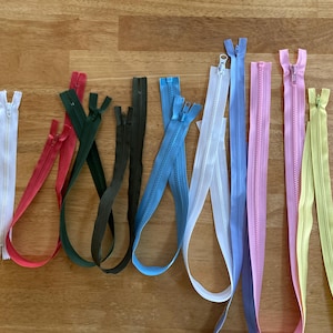 May include: A collection of colorful zippers in various shades, including white, red, green, black, blue, light blue, pink, and yellow. The zippers are arranged on a wooden surface, showcasing their different colors and lengths.