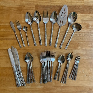 May include: A collection of stainless steel flatware featuring a bamboo-inspired handle design. The set includes various knives, forks, spoons, a slotted serving spoon, a solid serving spoon, a ladle, and a cake server, all with a polished finish.