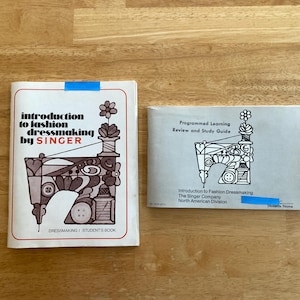 May include: Two vintage sewing books on a wooden surface. The book on the left is titled "Introduction to Fashion Dressmaking by SINGER" with a sewing machine illustration. The book on the right is a study guide.