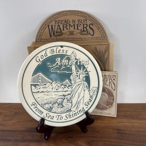 May include: A round ceramic bread warmer featuring a teal and cream illustration of the Statue of Liberty, a bridge, and wheat fields. The text reads God Bless America and From Sea To Shining Sea. It is displayed on a black stand with its original box.