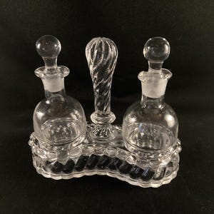 May include: A clear glass oil and vinegar set with three bottles and a twisted glass handle. The bottles have a textured surface and are set on a clear glass base.