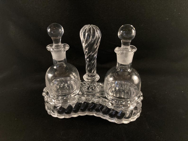 May include: A clear glass oil and vinegar set with three bottles and a twisted glass handle. The bottles have a textured surface and are set on a clear glass base.