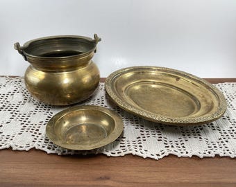 May include: Three antique brass items: a small pot with handles, a small round dish, and a large oval platter with a decorative rim. The items are arranged on a white lace doily and a dark wooden surface.