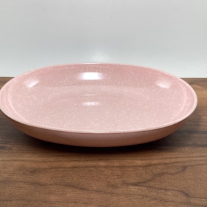 May include: A round, light pink serving bowl with a speckled pattern. The bowl is made of ceramic and has a wide, shallow design. The bowl is sitting on a dark brown wooden surface.