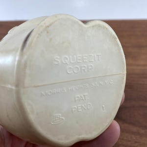 May include: Off-white plastic toy with the text "SQUEEZIT CORP" and "MORRIS HEIGHTS 53, N.Y.C." The bottom of the toy also has the text "PAT. PEND." and the number "1". The toy is held by a person.