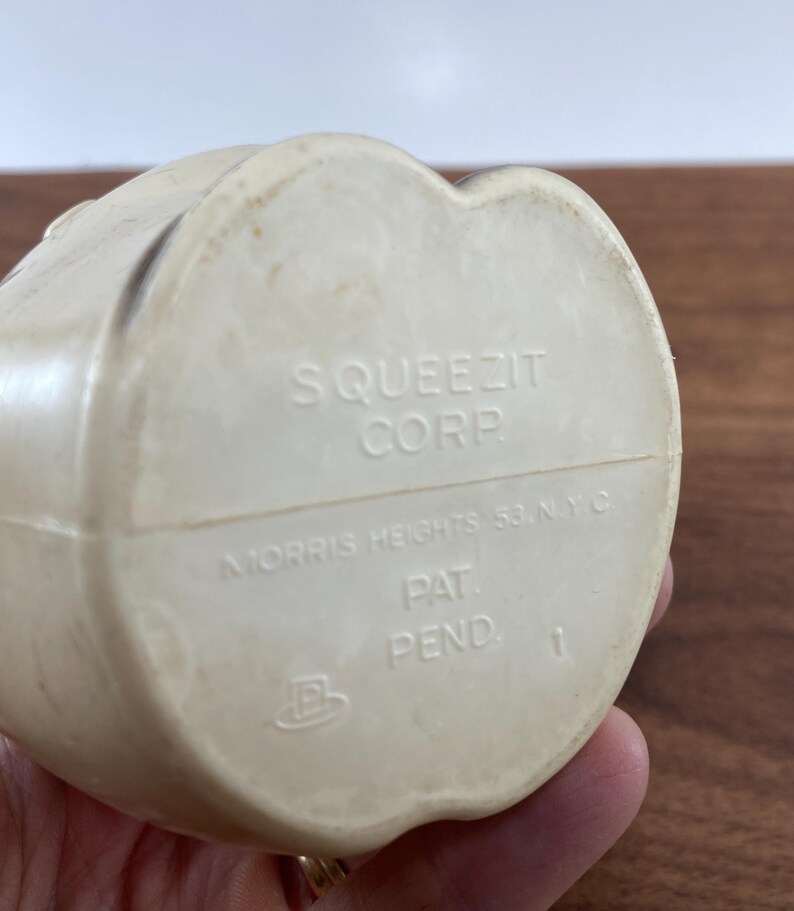 May include: Off-white plastic toy with the text "SQUEEZIT CORP" and "MORRIS HEIGHTS 53, N.Y.C." The bottom of the toy also has the text "PAT. PEND." and the number "1". The toy is held by a person.