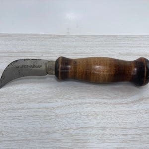 May include: A curved blade knife with a wooden handle. The blade is silver and has the text "JTA-SHARP" etched on it. The handle is made of wood with a dark brown finish.