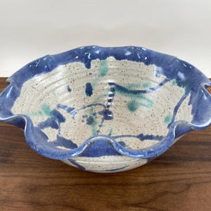May include: A ceramic bowl with a wavy, scalloped rim. The bowl features a speckled, off-white interior with blue and teal paint splatters. The rim is a solid, deep blue. A decorative piece for the home.