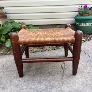 May include: A small footstool featuring four dark brown wooden legs with rounded tops and a woven tan rush seat. The stool has a rectangular frame with horizontal support bars connecting the legs, providing a stable base for the woven surface.
