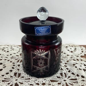 May include: A ruby red crystal jar with a lid and a clear glass knob. The jar has intricate cut glass designs, including stars and floral patterns. The label reads "Crystal Legends by Godinger, Handcrafted Crystal, Made in Romania."