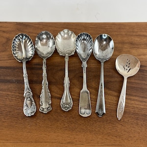 May include: A collection of six silver-toned metal spoons arranged in a row. The set includes five ornate spoons with varied bowl shapes and decorative handles, and one smaller, round-bowled spoon featuring a pierced wheat-like pattern design.