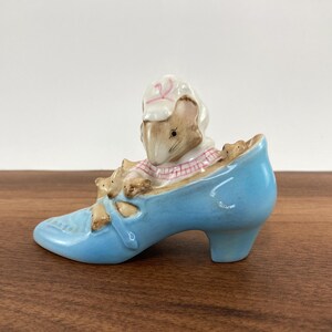 May include: A ceramic figurine of a mouse wearing a bonnet and a plaid shirt, sitting inside a light blue high-heeled shoe. The shoe has a decorative bow on the front. The figurine is set on a wooden surface.