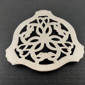 May include: A silver-toned metal trivet featuring an intricate, symmetrical openwork design with curved, interlocking patterns. The object has a wide, shield-like shape with a smooth, solid border and a detailed, cut-out center section.