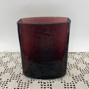 May include: A square-shaped, dark red glass vase with a textured surface. The vase has a gradient effect, transitioning from a lighter red at the top to a deep, almost black color at the base. The interior is a lighter shade of red.