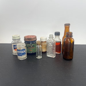 May include: A collection of eight vintage glass bottles and jars of varying sizes and colors, including clear, amber, and blue glass. Several containers feature original paper labels with text such as Bufferin, Vicks, and Holy Water.
