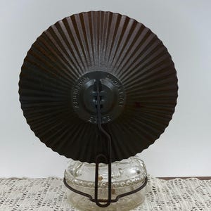 May include: A vintage, dark brown metal plate with a radial design, attached to a clear glass jar with a metal handle. The plate has the text "MADE AT HOME" and "A.B.U. IN 30AM". The jar sits on a white lace doily.