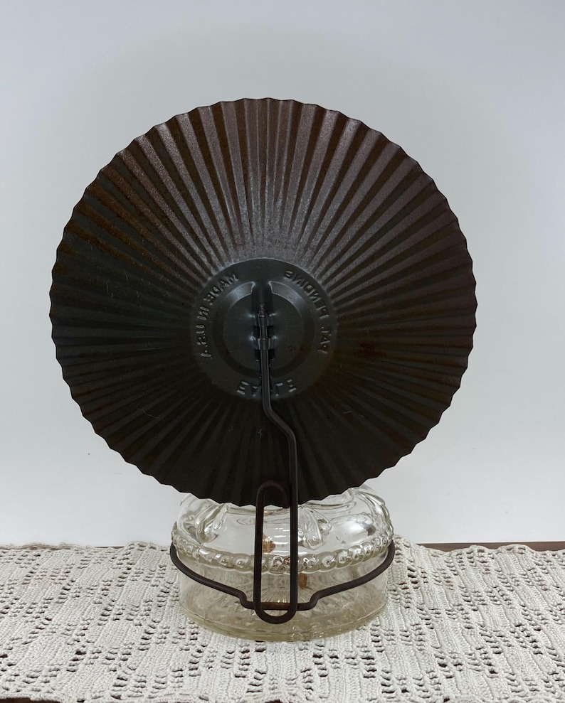 May include: A vintage, dark brown metal plate with a radial design, attached to a clear glass jar with a metal handle. The plate has the text "MADE AT HOME" and "A.B.U. IN 30AM". The jar sits on a white lace doily.