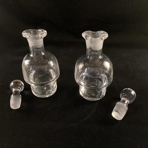 May include: Two clear glass bottles with stoppers, each with a textured, rounded base. The bottles are empty and have a slightly frosted appearance.