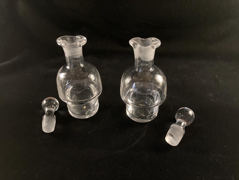 May include: Two clear glass bottles with stoppers, each with a textured, rounded base. The bottles are empty and have a slightly frosted appearance.