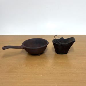 May include: A small, dark, cast iron skillet with a single handle and three short legs, positioned next to a small, dark, cast iron bucket featuring a thin metal wire handle and a wide, open top with a slightly curved rim.