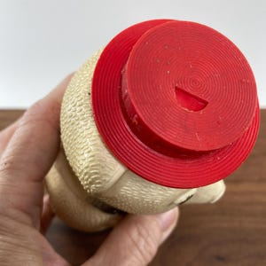 May include: A vintage, cream-colored rubber toy with a red hat. The toy has a textured surface and a small opening on the hat. The toy is held in a hand.