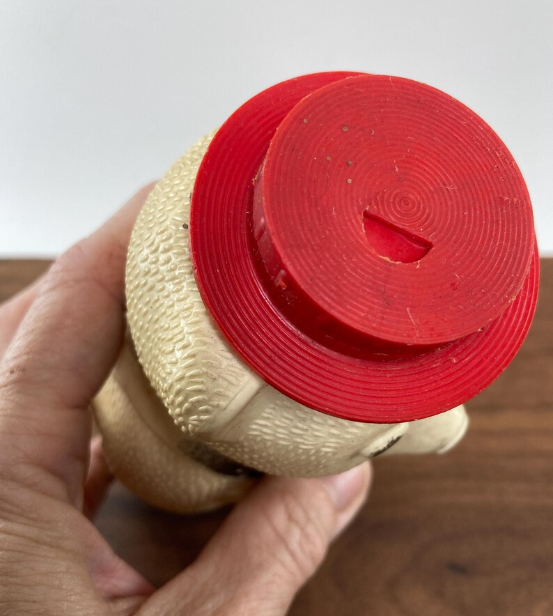 May include: A vintage, cream-colored rubber toy with a red hat. The toy has a textured surface and a small opening on the hat. The toy is held in a hand.