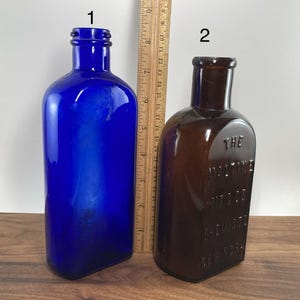 May include: Two antique glass bottles. One is a vibrant cobalt blue, the other is brown. The brown bottle has embossed text that reads "THE MALTINE MFG. CO. CHEMISTS NEW YORK". A wooden ruler is placed between the bottles.